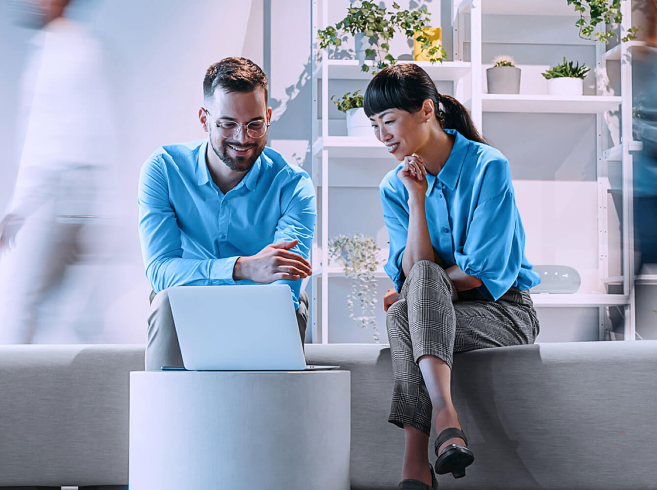 Zwei Personen sitzen auf einer Couch und blicken auf einen Laptop auf einem kleinen runden Tisch, im Hintergrund sind weitere Personen in Bewegung und ein Regal mit Pflanzen zu sehen.